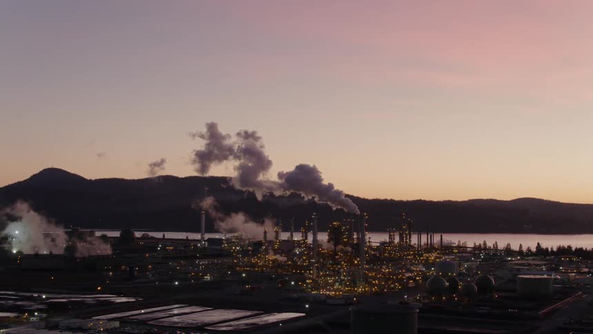 Smoke and steam billowing from refinery smokestacks below a beautiful sunset sky