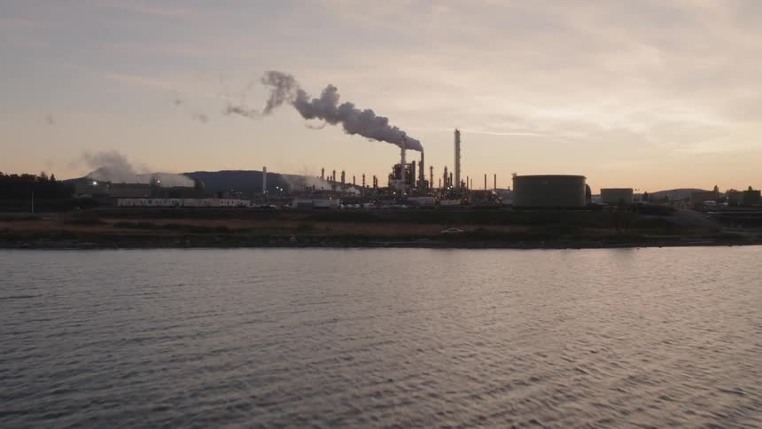 Skimming low over calm harbor water toward an oil refinery