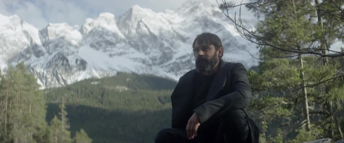 A bearded man sits out in nature by a snow covered mountain