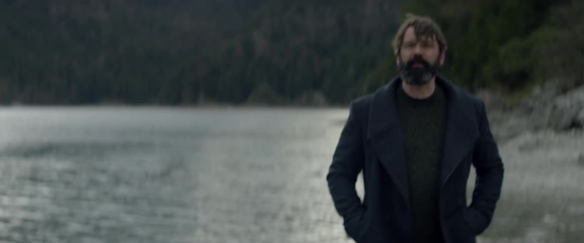 A bearded man stands by the water and talks