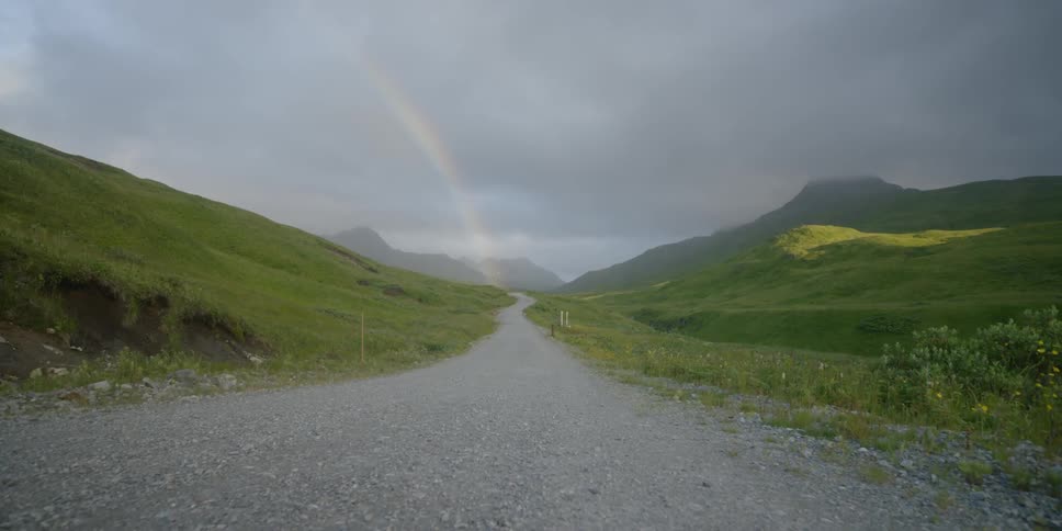 A rainbow is shining over a road in a natural setting on a stormy day
