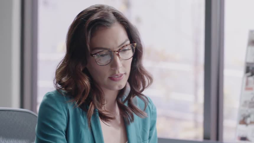 Woman places on eyeglasses as she's working at a desk indoors and then removes them