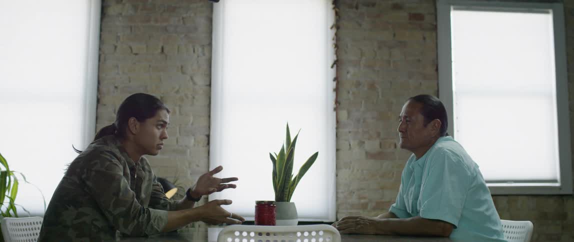 Two men talk at a table near windows