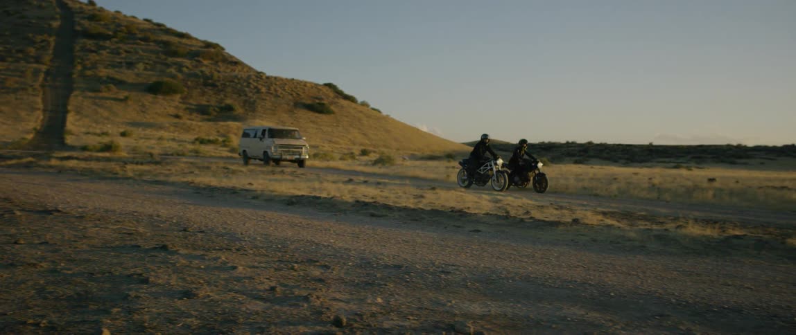 A van follows two motorcycle riders on a dirt road at sunrise