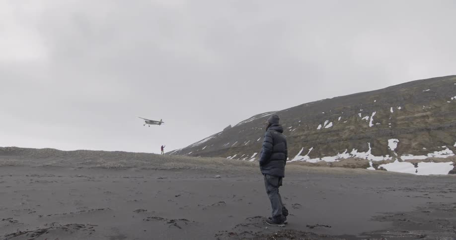 A man in a coat stands on the beach as a plane flies overhead