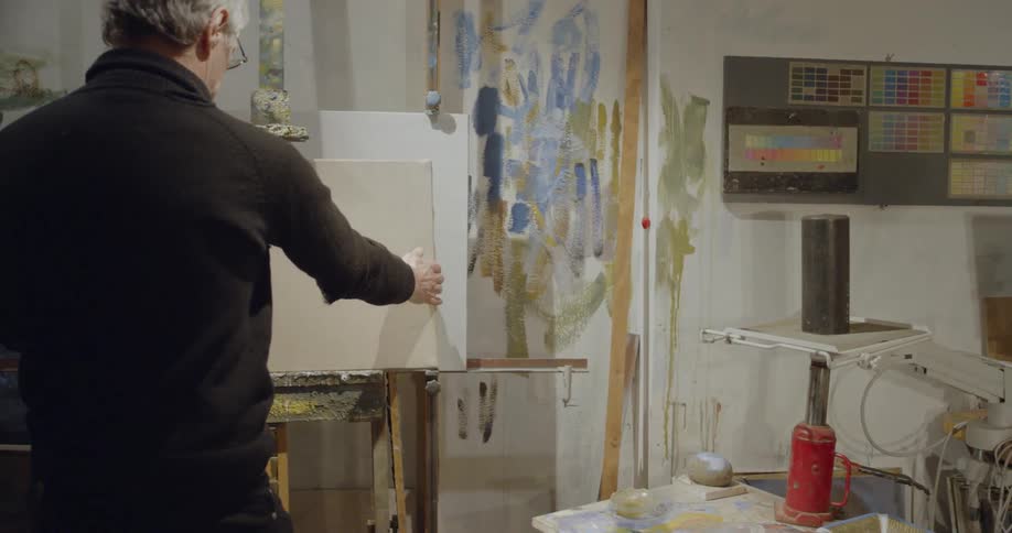 A man picks up a blank canvas and places it on an easel