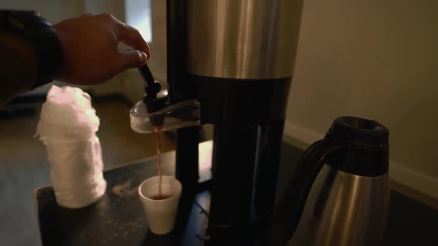 A person pours a cup of coffee from an urn on a table