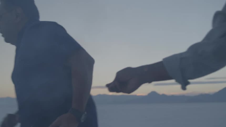 Two men hand off a book while running in salt flats at sunset