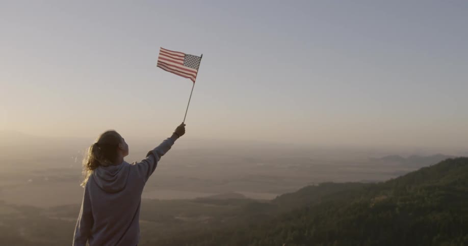 Girl holds up an american flag while overlooking a valley at sunset
