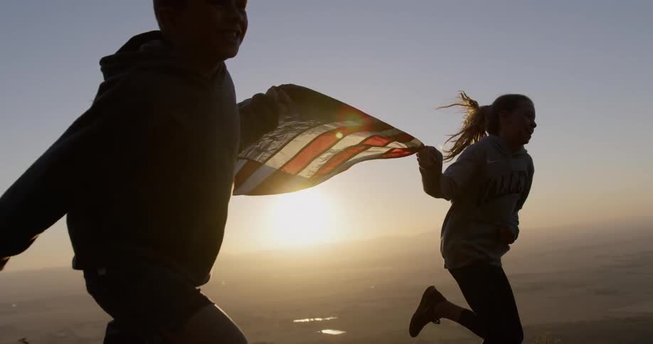A boy and girl holding a us flag are running in a natural setting at sunset