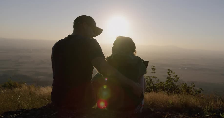 Father and daughter sit on hill top watching sunset