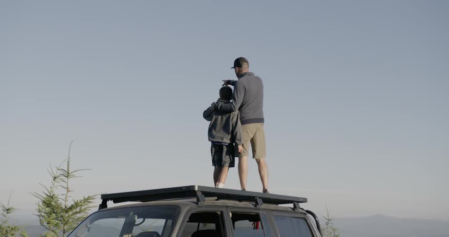 A man and a kid are standing on the roof of their car looking out over the edge of a cliff