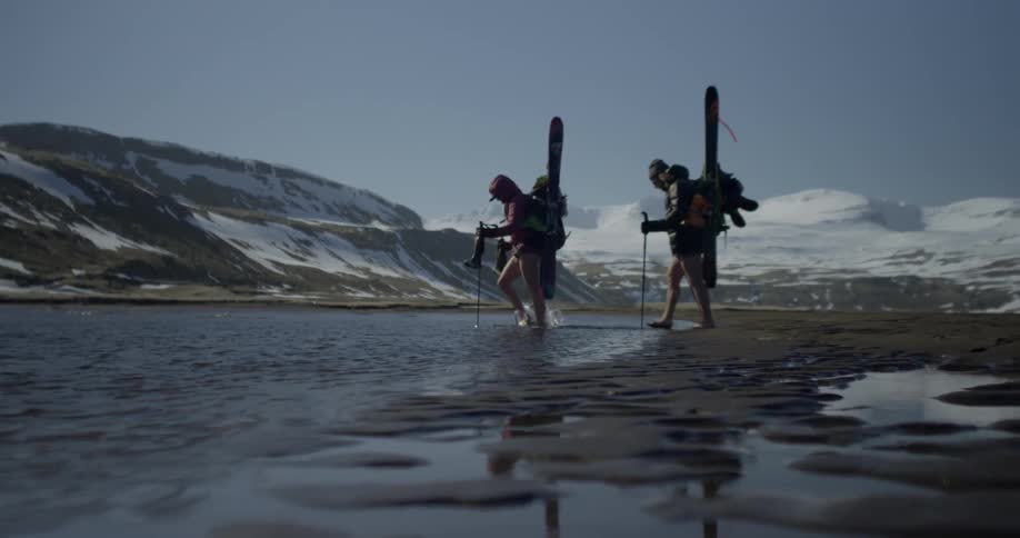 Hikers carrying skis are wading across a river during the day