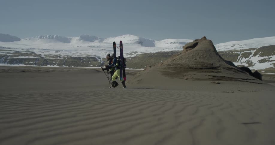 Two hikers carrying skis are trudging across a sandy area during the day