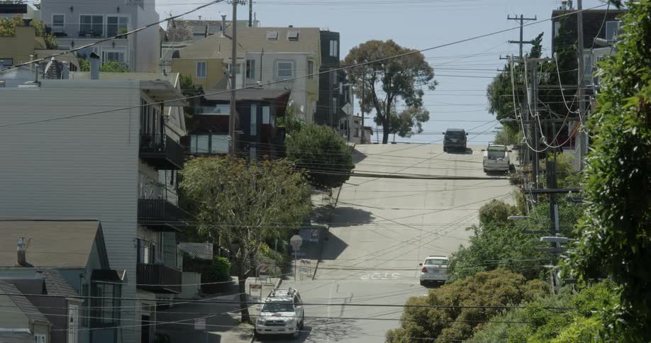 Vehicles are traveling up a steep street in a city during the day