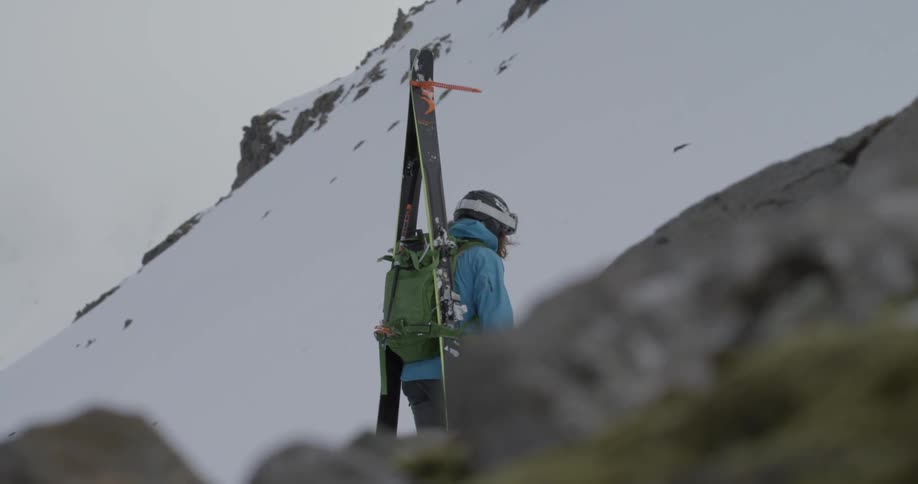A person carries a backpack and skis as they hike up a mountain during the day