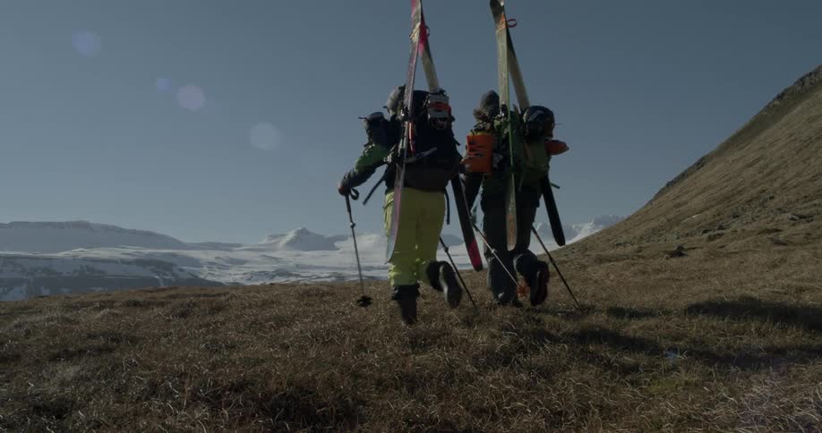 Two hikers carrying their skis walk through the grass toward snow