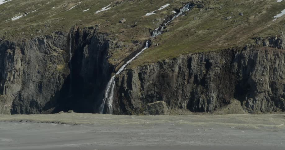 A distant waterfall flows down a grass hillside onto the beach