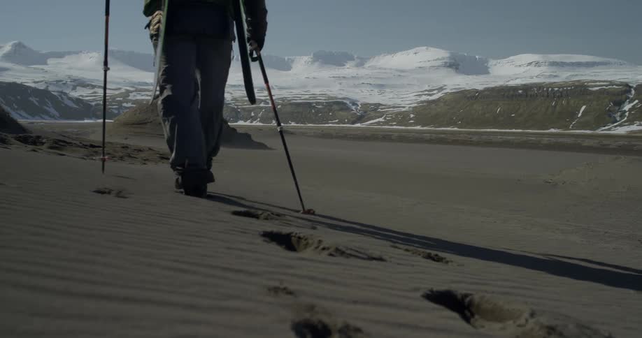 Man walking across a barren area with ski poles and mountains in the distance