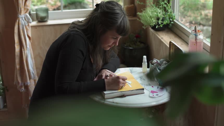 A woman is painting in a book as she sits at a table during the day