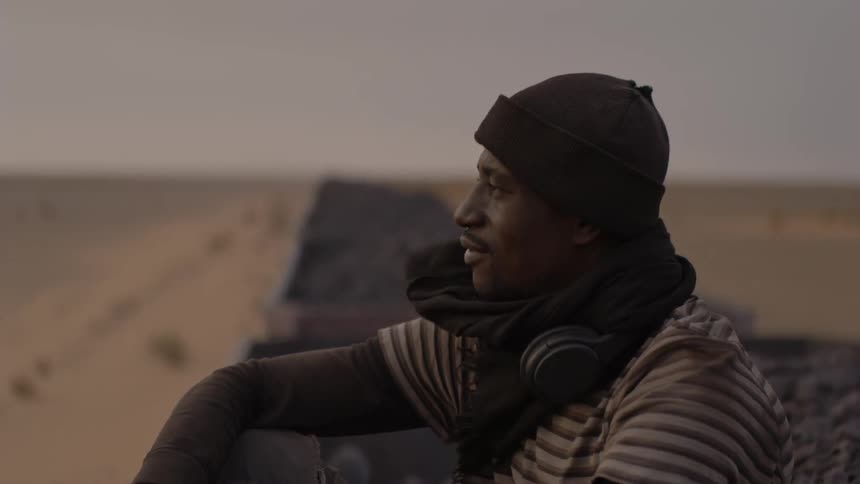 A man sits on top of a rail car moving through the desert