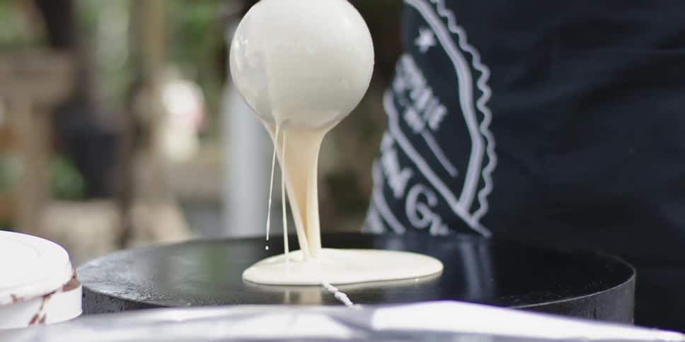 A cook is pouring crepe batter onto a hot plate outside during the day