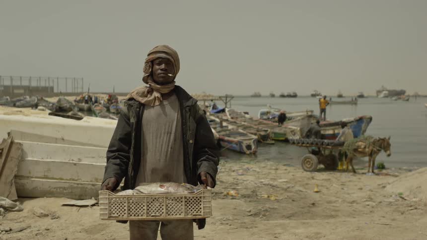 A man with a cigarette in his mouth stands on the sand holding a crate