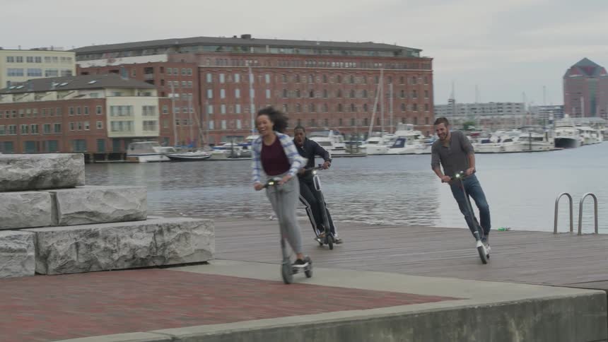 Friends are riding scooters along a boardwalk in a city on a cloudy day