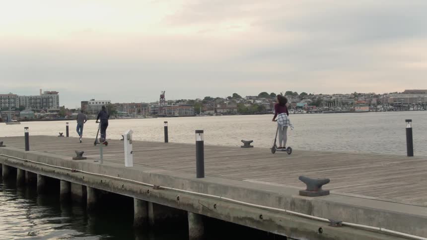 Friends are riding scooters along a pier in a city at dusk