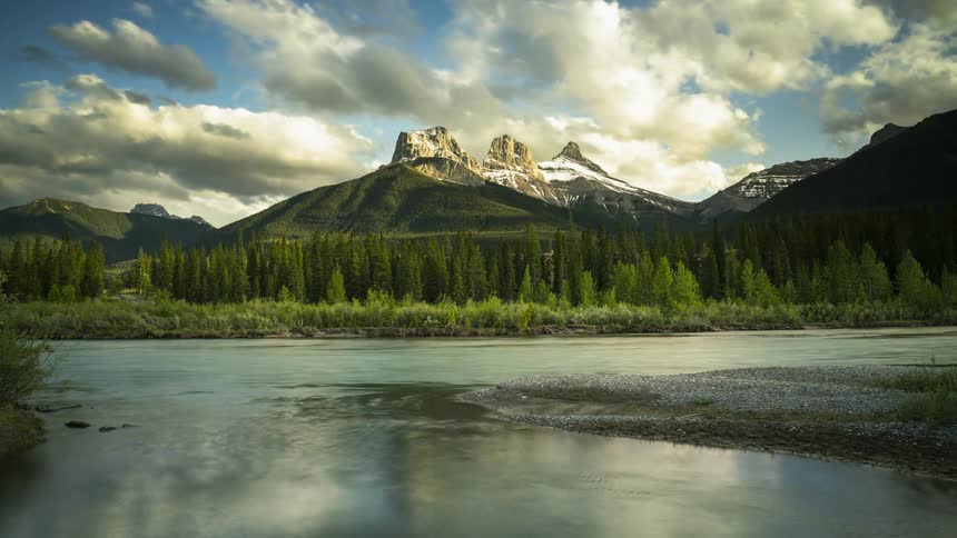 The Three Sisters and clouds overhead are reflected in the Bow River in Banff, Canada