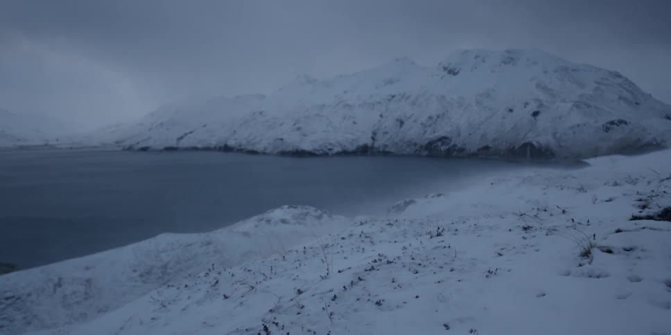 The wind blows snow from the hills on shore on a cloudy day