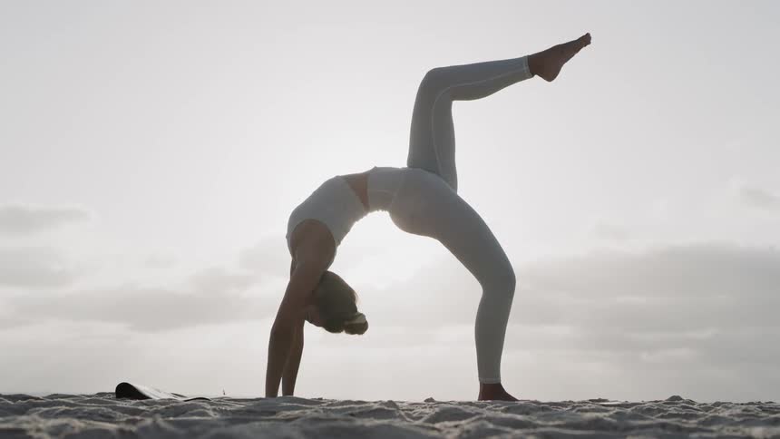 A woman is doing a backbend on the beach and then extends one leg into the air