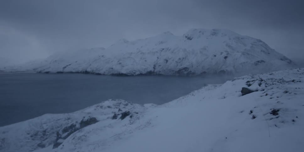 The wind drifts snow across the hill towards the water