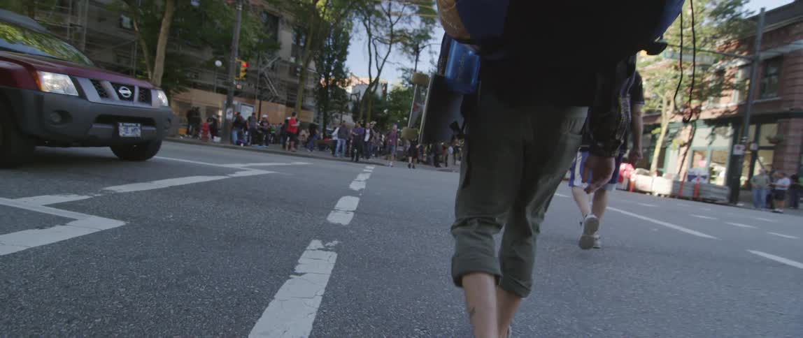 Man walking with a skateboard down a street towards a large group of people