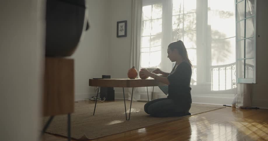 A woman is sitting on the ground in her home carving pumpkins during the day