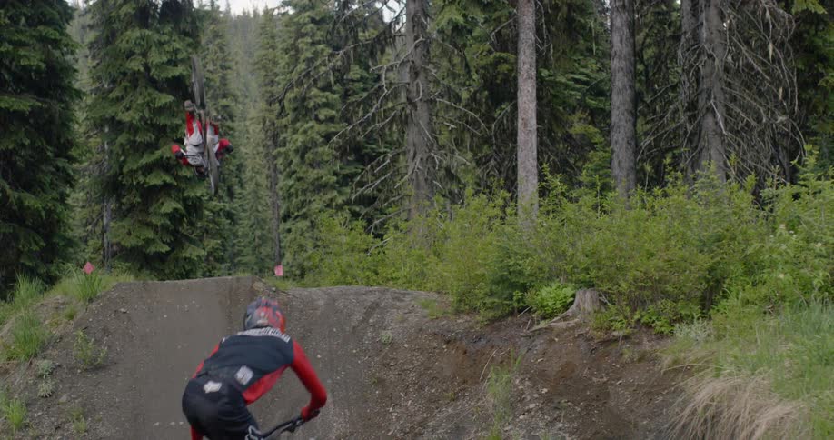 A man is racing a mountain bike up a ramp on a trail and performing a stunt