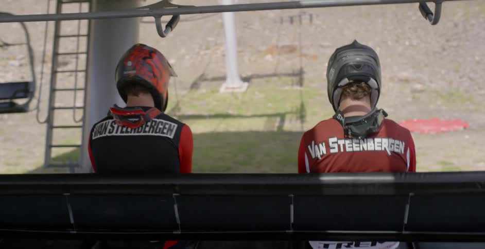Two people ride a ski lift while wearing motocross gear