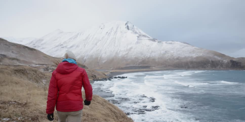 A man in a red coat walks on the rocky shore