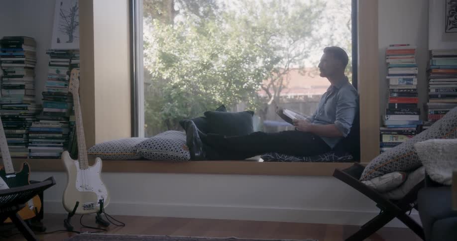 A man is sitting in a window reading a book during the day