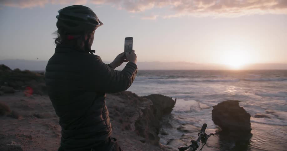 A bicyclists is taking pictures of a coastal view at sunset