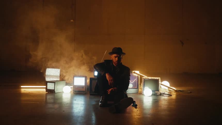 Man sitting in an area with many monitors behind him including one that appears to be smoking