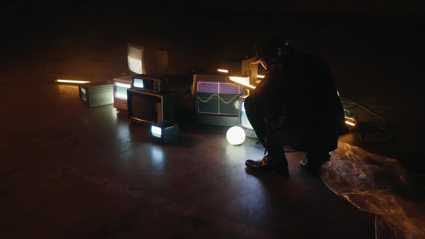 A man is squatting and looking at a collection of monitors on the floor in a room