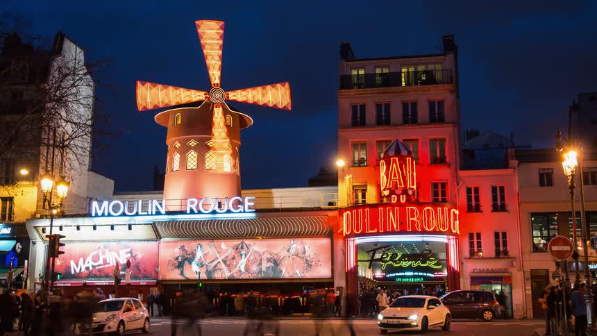 Pedestrians and traffic are passing in front of the moulin rouge at night