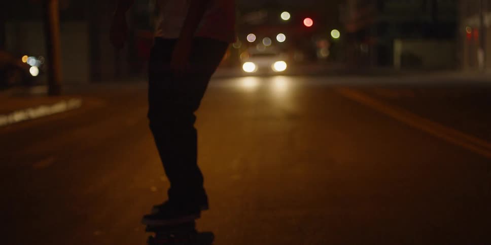 Teenager pushes a skateboard down a city road at night with headlights behind