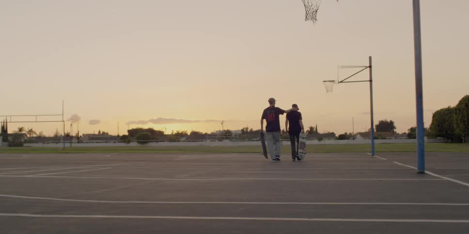 Two teenagers are carrying skateboards across a basketball court outdoors at dusk