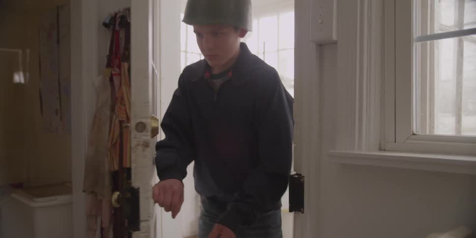 A boy wearing a toy army helmet walks into a house