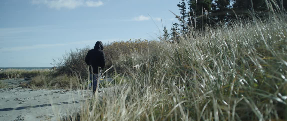 A man in a hoodie is walking on the beach towards some tall grass