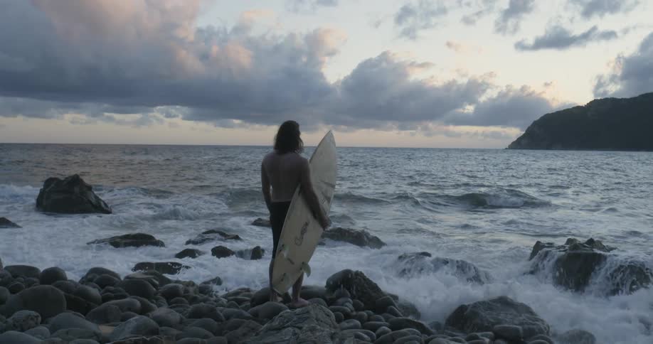 A surfer stands on a rocky beach at sunset