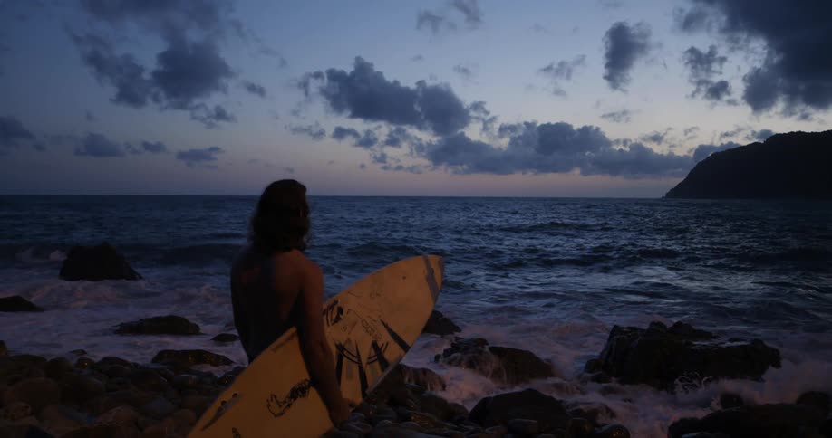 A man holds his surfboard and looks out at the ocean at sunset