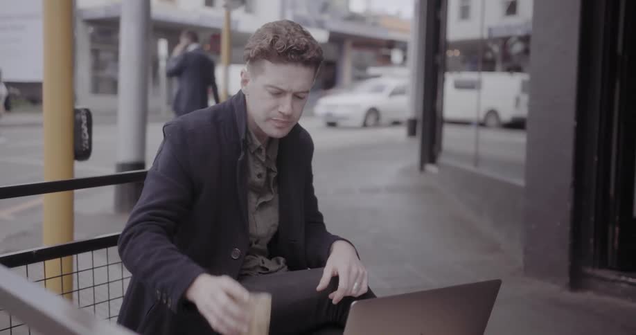 Man drinks coffee while sitting outside typing on a laptop